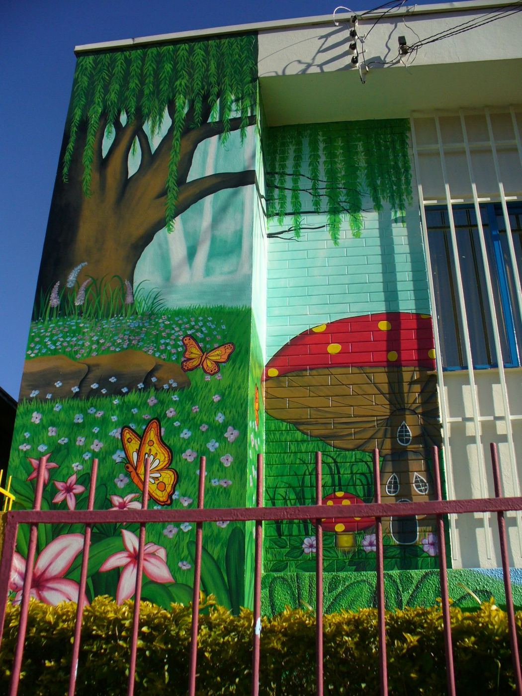 Fachada de prédio pintada com mural de jardim: árvore, cogumelo vermelho, flores e borboletas